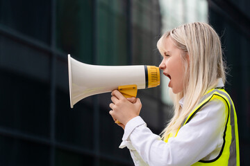 Young woman activist wearing reflective safety vest stands outdoors and speaking into a megaphone ...