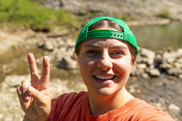 Cheerful young woman in a green cap and orange shirt smiling and showing a peace sign during a hike...