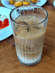 A glass of iced milk coffee with a clear coating, placed on a wooden table, with a slightly blurred background of food dishes, outdoor cafe.