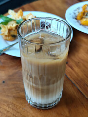 A glass of iced milk coffee with a clear coating, placed on a wooden table, with a slightly blurred background of food dishes, outdoor cafe.