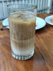 A glass of iced milk coffee with a clear coating, placed on a wooden table, with a slightly blurred background of food dishes, outdoor cafe.