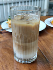 A glass of iced milk coffee with a clear coating, placed on a wooden table, with a slightly blurred background of food dishes, outdoor cafe.