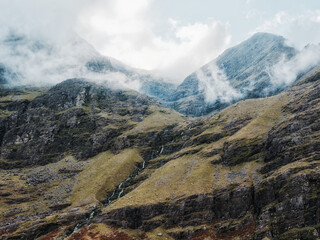 Mountainside reveals cascading water flowing down rocky terrain. Sparse vegetation clings to the slopes. Clouds partially obscure the peaks.