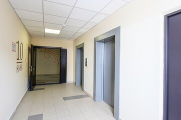 well-lit hallway with tiled floors, a drop ceiling, two elevators, and an open doorway with apartment numbers labeled on the wall