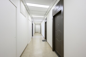 long, well-lit corridor with tiled floor and white walls. Several dark wood doors are visible. Modern ceiling with fluorescent lighting