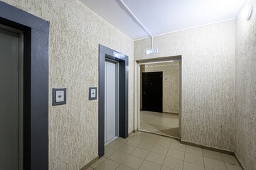 Interior shot of a hallway with beige walls, a tiled floor, and doorways. Two elevators are on the left, and a doorway leads to another room with a black door. The walls have a textured finish