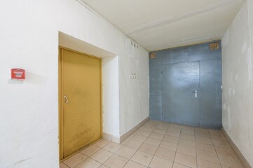interior shot of a hallway featuring a yellow door, a grey metal door, and tiled flooring. Walls are white, showing signs of wear
