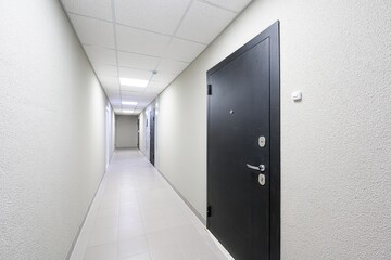 long, well-lit hallway with white walls, tile floors, and dark doors on either side. The ceiling is a drop ceiling with square panels and recessed lighting