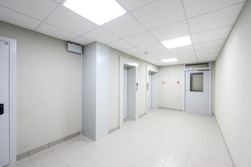 bright corridor with two elevators, tiled floor, and a door. The walls are off-white, and the ceiling has square light panels