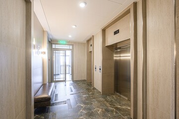 modern lobby with elevators, marble floors, and neutral tones. There's a bench, exit sign, and good lighting. Clean and inviting