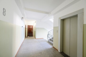 image shows an indoor view of an apartment building hallway, featuring an elevator, staircase, and entrance doors. The walls are painted in a two-tone scheme of light green and white