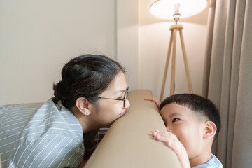 Asian mother with glasses and her children playing peekaboo and laughing behind sofa in living room.