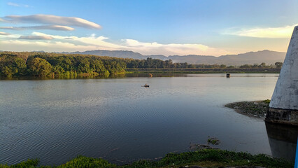 A truly beautiful morning view. The sky is so clear and blue. The reservoir is set against a backdrop of hills and mountains. Someone is fishing in a boat.