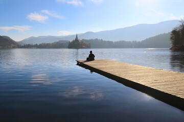 Man sitting on the pier of Lake Bled looking towards the Assumption Church island.