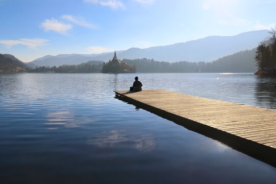 Man sitting on the pier of Lake Bled looking towards the Assumption Church island.