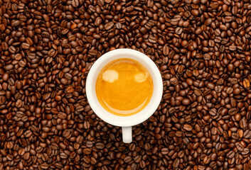Top view of a white cup filled with fresh espresso on blurred roasted coffee beans background. Perfect aroma, crema texture, and warm brown tones create a cozy and energetic mood.