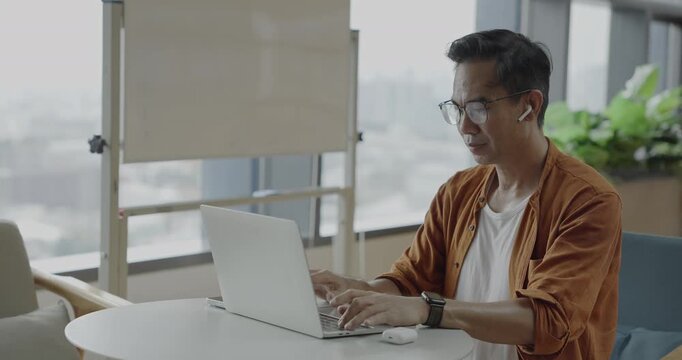 Senior Asian businessman wearing headset celebrates success during online meeting in modern co-working space. Remote work and business communication concept.