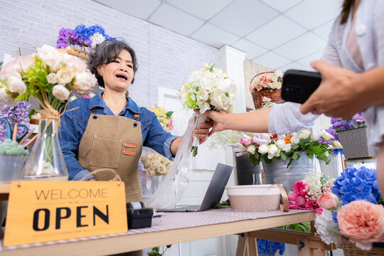 Small business lifestyle, senior working woman handing rose bouquet to customer for Valentine's Day, silver-haired Asian elderly shop owner engaging warmly with client in flower store focus open sign - Powered by Adobe