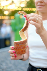 Woman with bright pink nails holding a spiral cinnamon-sugar coated trdelnik pastry with a glowing outdoor festival sunset behind, capturing street food joy and warm atmosphere