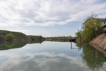 Obraz premium Calm lake with reflections, wooden pier, cloudy sky, peaceful nature scene, serene water, green landscape, rural area, fishing spot, outdoor relaxation, tranquil environment.