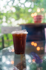 A plastic cup filled with iced black coffee sits on a glass table with a blurred background.