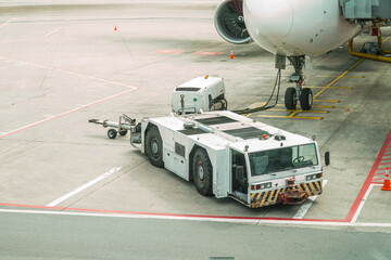 Aircraft tug is parked near the airplane ready for operation. Aviation ground support equipment for pushback. Transportation hub with modern plane and tow tractor. Preparing for travel.