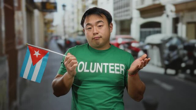 Young man holding cuban flag with unsure expression on city street wearing volunteer shirt