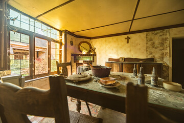 Abandoned rustic farmhouse dining room with dusty tableware, old bottles, and vintage decor illuminated by natural light through large windows