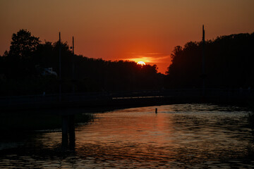 Fototapeta premium Sunset over calm river with dark bridge silhouette and trees reflecting warm evening sky colors in tranquil water creating serene scenic atmosphere