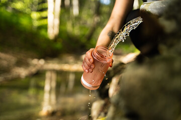 Close-up of a person filling a reusable plastic bottle with clean spring water in the mountain...