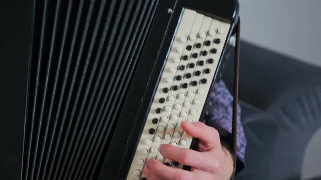 Extreme close-up of a young bard hand as he presses the accordion buttons with care. The male singer-songwriter, wearing glasses and a patterned shirt, listens to the sound while selecting a melody. 