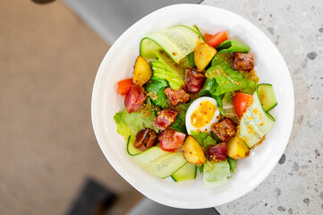 Fresh salad with lettuce, cucumber, tomato, grilled meat, boiled egg, peach slices, and mustard dressing served in a white bowl on a light background.
