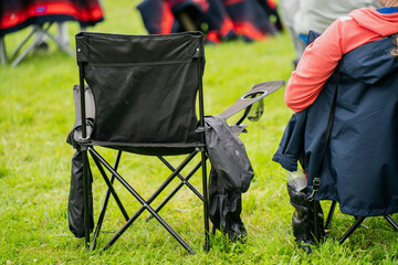 Black folding camping chair on green grass at outdoor event with people in background, used for festivals, concerts, sports or leisure activities in summer open-air setting