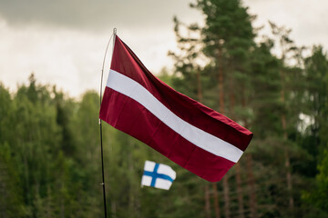 Latvian and Finnish flags waving in outdoor setting with forest background, national flags flying together at international event or sports competition in natural environment