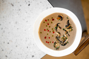 Bowl of creamy mushroom soup with sautéed mushrooms, green herbs, and colorful sauce dots, served on a speckled countertop, top view.
