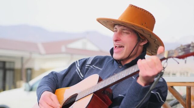Romantic man in a cowboy hat sings with emotion and plays guitar while sitting outside in soft daylight. His eyes gently squint as he enjoys music, evoking feelings of love and connection to nature