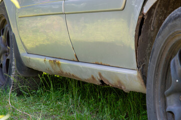 A car with visible rust and deterioration is partially hidden in tall grass, showcasing signs of neglect in a outdoor environment
