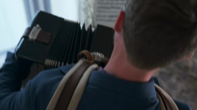 Overhead shot of a man in formal outfit recording a Bayan accordion lesson at home for an online learning platform. Sheet music is placed on a stand in front of him.