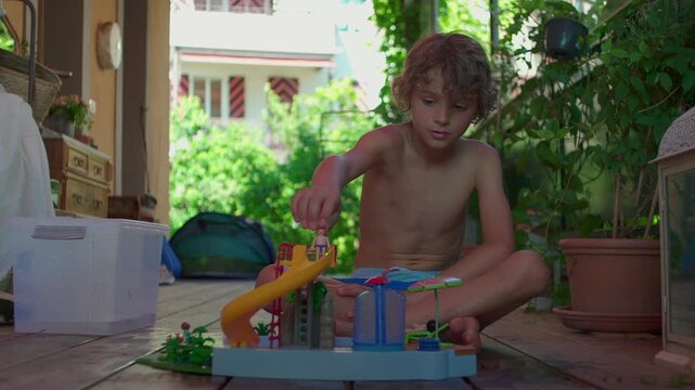 Child plays alone with colorful plastic water slide toy on wooden patio, focused and seated in peaceful garden space on sunny day