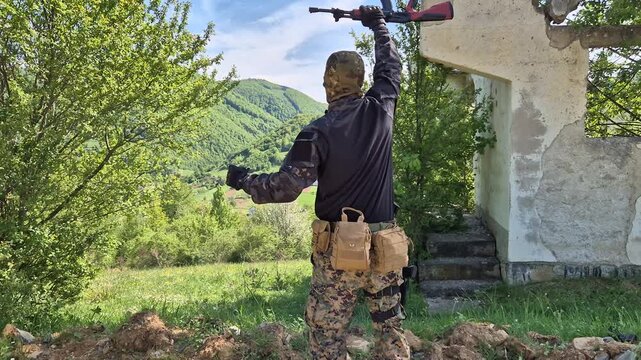 Armed man terrorist combatant militant soldier in forested hills, holding rifle over shoulder overlooking a village city in combat
