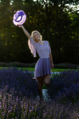 A woman in a flowing lavender dress joyously lifts a hat against the backdrop of a lush lavender field, embodying the essence of a sunny day in nature. The scene is beautifully lit,