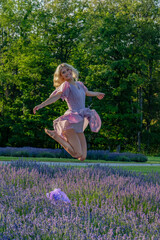 A woman in a pink dress joyfully leaps in the air above a field of blooming lavender flowers. The...