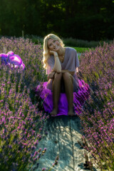 A woman sits on a path surrounded by blooming lavender fields, her light dress and the gentle sunlight creating a serene atmosphere. She smiles warmly, capturing a moment of tranquility 