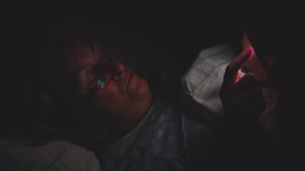 Older woman lying in bed at night using smartphone, face illuminated by screen light in dark bedroom, wearing glasses and focused on device - Powered by Adobe