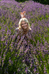A blonde woman with a joyful expression lies among lush fields of blooming purple lavender. Sunlight bathes the scene, highlighting the vibrant colors and serene atmosphere.