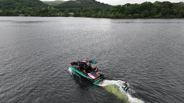 Aerial side angle tracking wake surfer and boat traveling down Ullswater lake Lake District UK cloudy day
