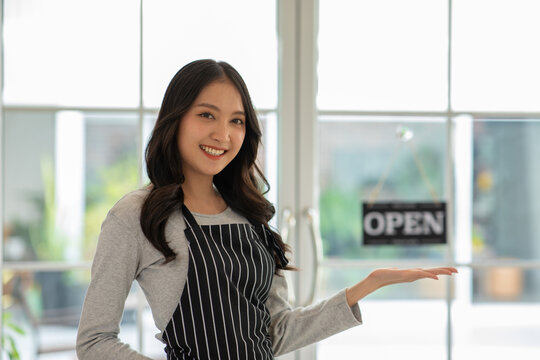 Asian Smiling woman wearing striped apron standing indoors near glass door with open sign, welcoming customers with friendly gesture and positive expression