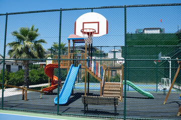 Basketball Court With Playground Equipment Featuring Slides and Climbing Structures During a Sunny...