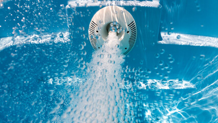 Close-up underwater view of a pool jet releasing bubbles with intense pressure, surrounded by blue water and light reflections.