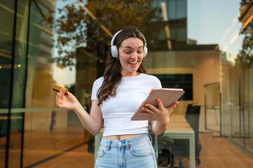 Excited young woman making online payment using tablet and credit card, listening music with headphones